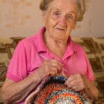 An old woman sits knitting a rug.